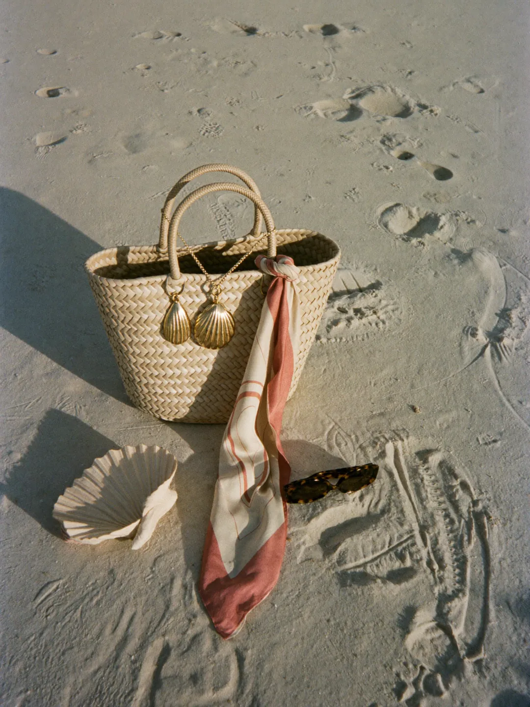 Straw tote bag, shell jewellery and a silk scarf arranged on pale sand